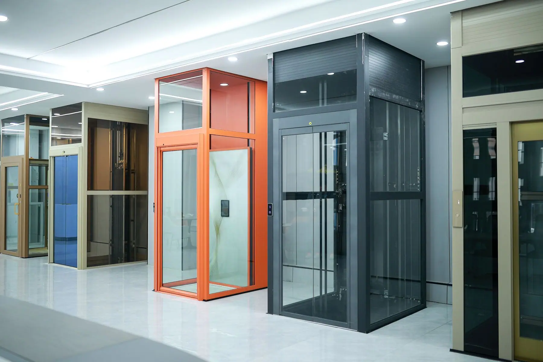Modern glass home elevator models in a showroom featuring orange and black frames for residential accessibility.