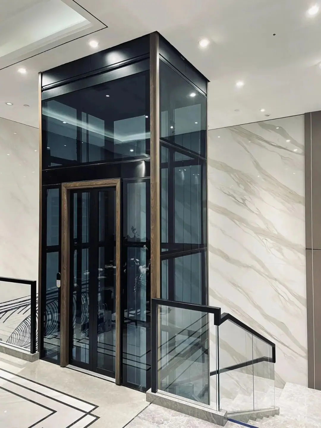 Levitex Max glass lift with a dark frame and bronze door trim beside a marble wall.